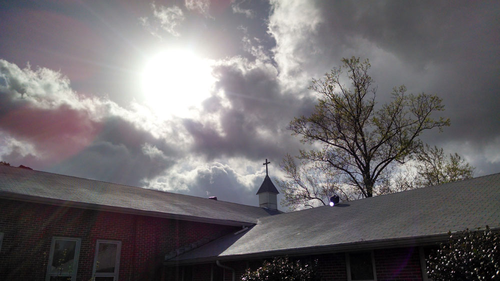Photo of sun breaking through clouds over monastery chapel steeple.